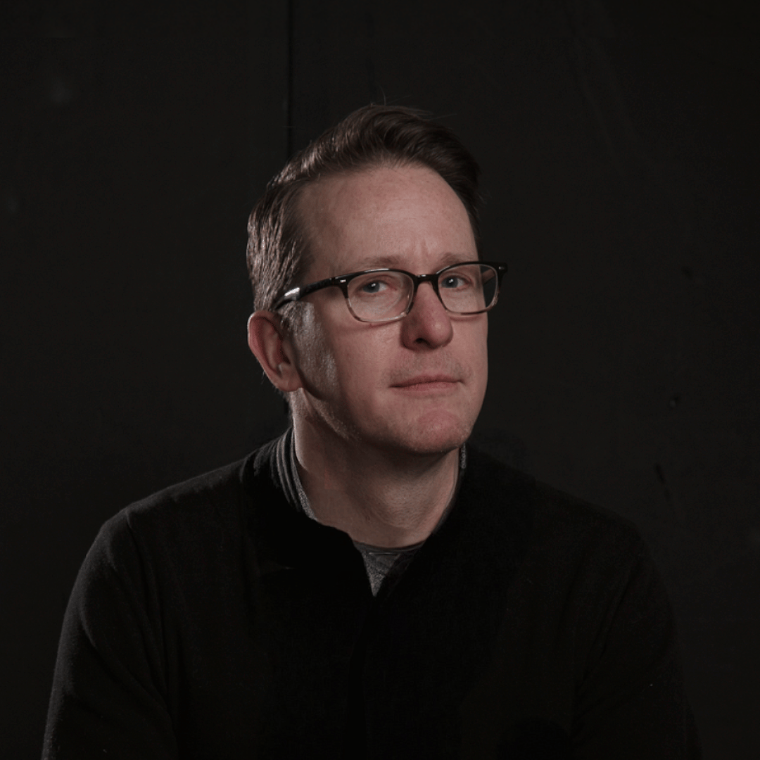 Portrait of a man with brown hair and glasses on a flat gray background.